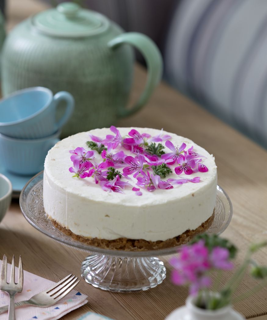 Violette Blüten eine Duftgeranie auf Kuchen. Im Hintergrund sind Besteck und Teekanne.