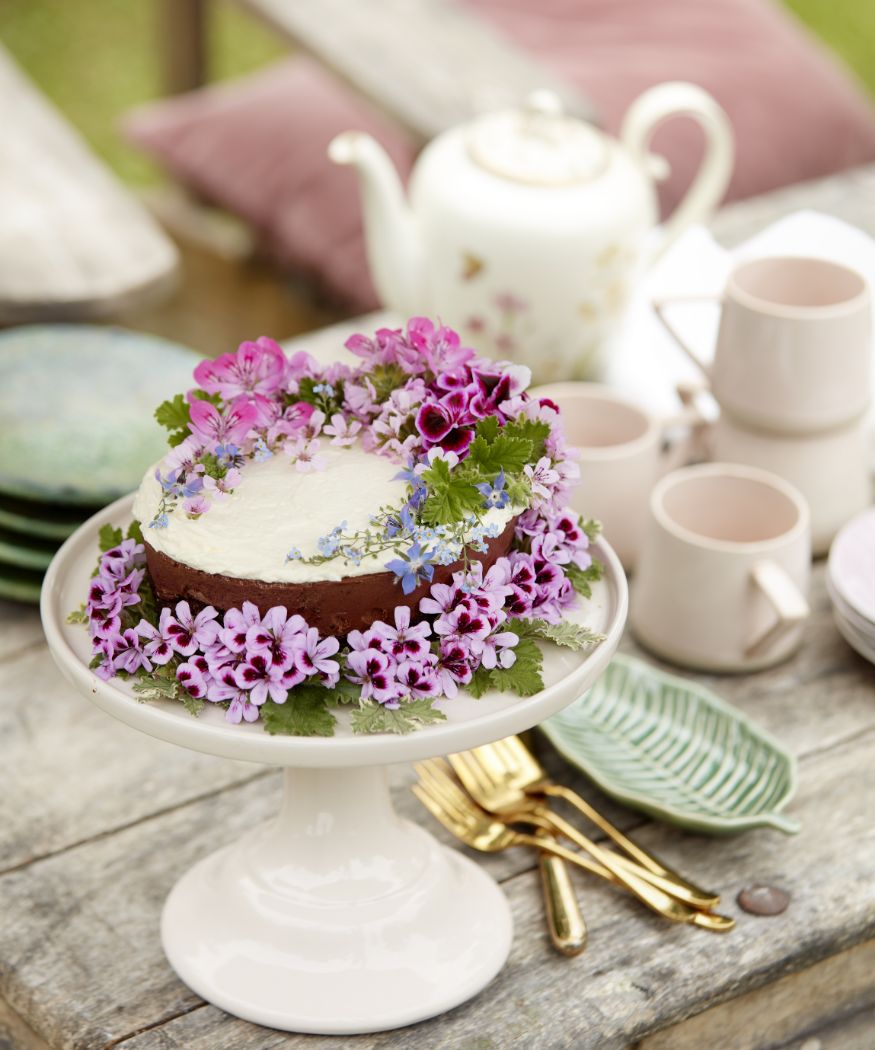 Kuchen dekoriert mit violetten Duftgeranien neben goldenem Besteck, Tellern, Tassen und Teekanne